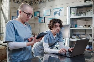 Two veterinary staff members in blue scrubs working together in a clinic; one uses a handheld tablet while the other types on a laptop near medical equipment.