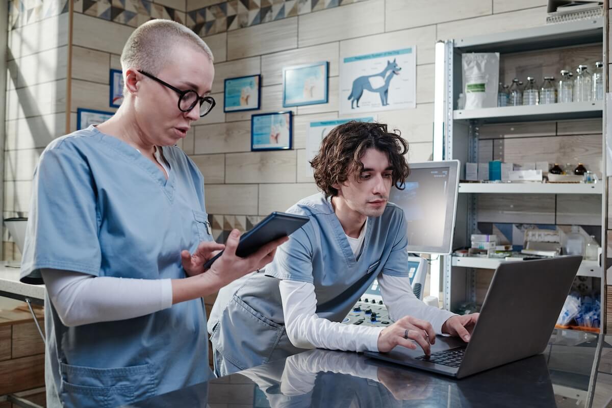 Two veterinary staff members in blue scrubs working together in a clinic; one uses a handheld tablet while the other types on a laptop near medical equipment.