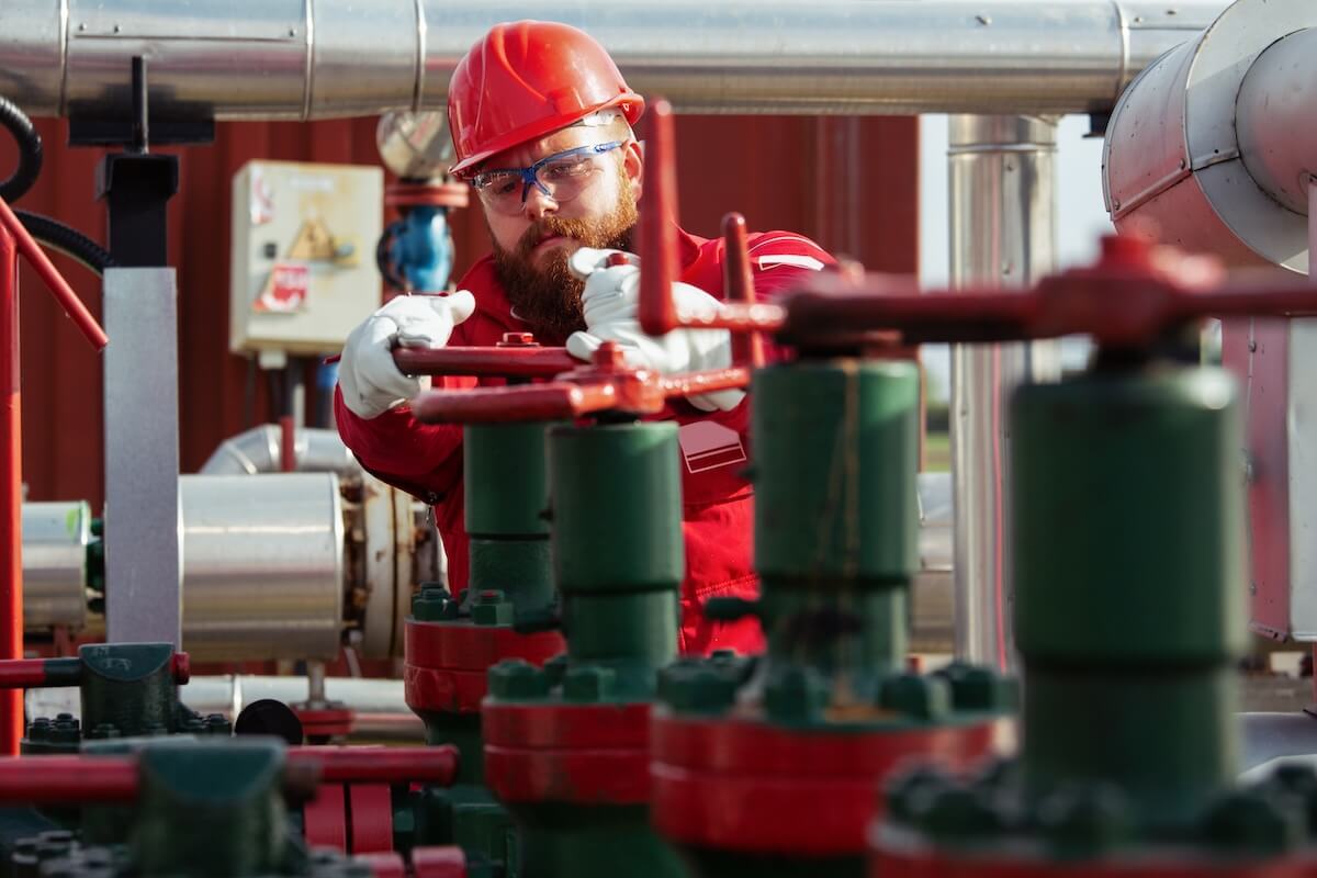 Oil worker turning valve on oil Worker in a red hard hat and safety gear turning a large valve on industrial piping.