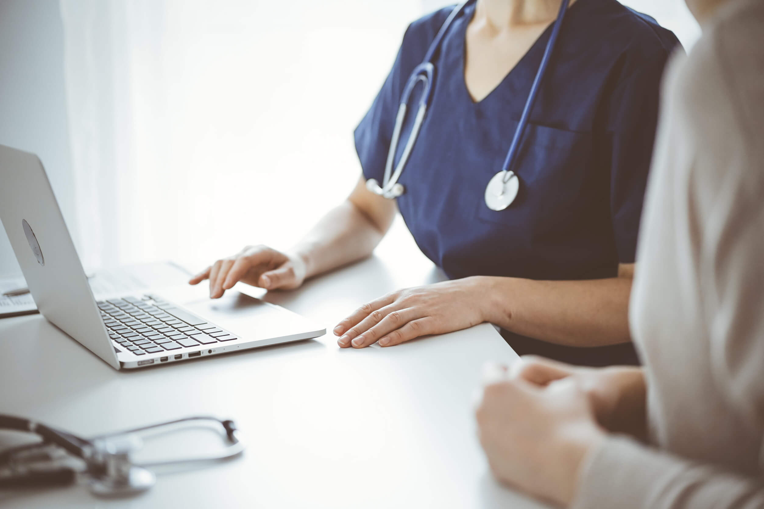 A close-up of a veterinary professional in blue scrubs using a laptop during a consultation at a white desk, representing the collaborative discovery phase of switching IT providers.