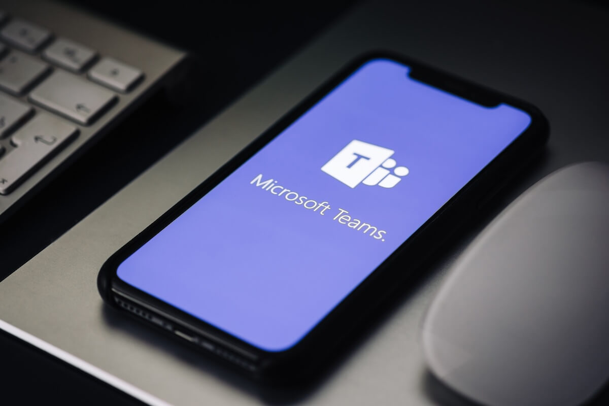 A smartphone on a desk displaying the Microsoft Teams logo next to a computer keyboard and mouse.