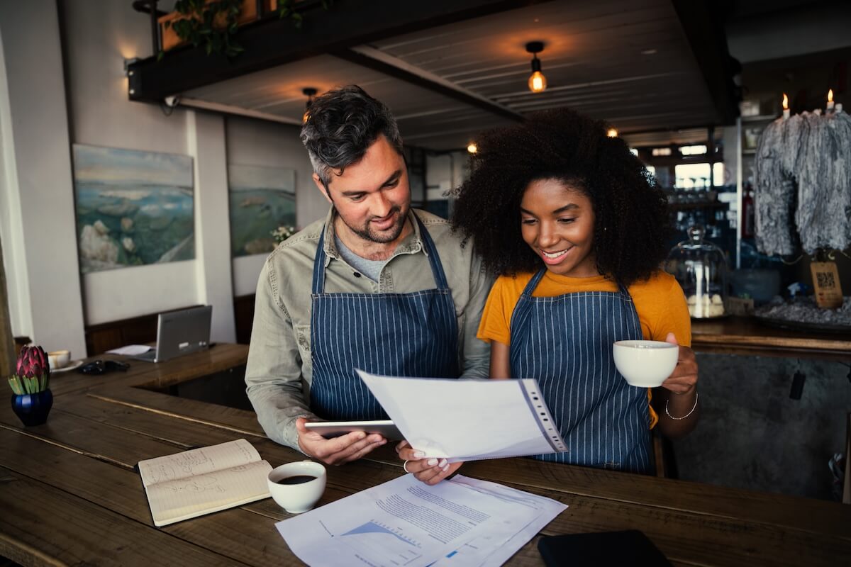 Man and woman in aprons reviewing a business data report and tablet at a cafe counter.