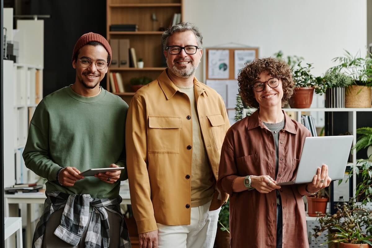 Three smiling small business professionals holding a laptop and tablet in a modern office with plants.