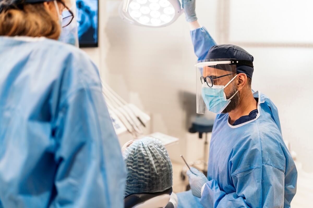 A dentist in a face mask adjusts an overhead light while treating a patient in a dental chair.