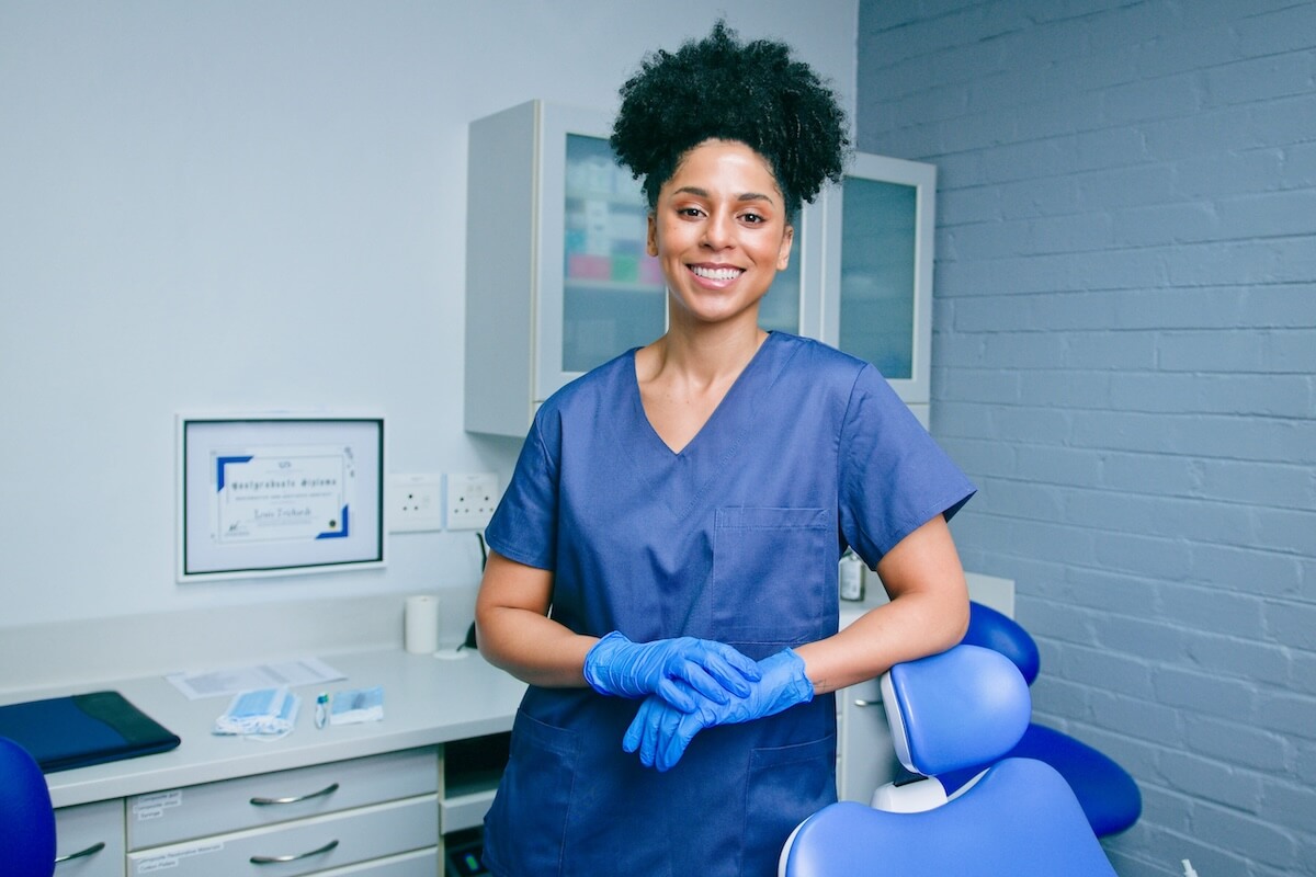 Smiling female dental professional wearing blue scrubs and gloves leaning on a clinical chair.