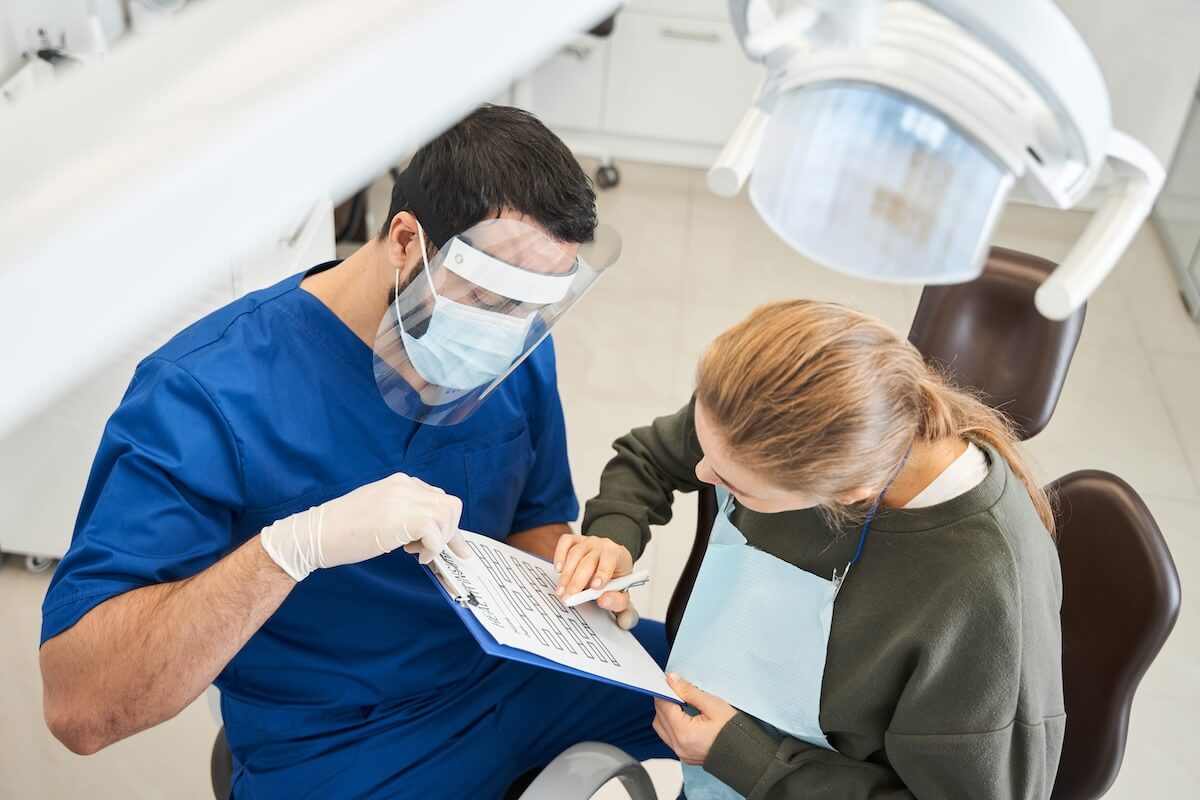 Overhead view of a male dentist wearing a face shield reviewing a clipboard form with a female patient.