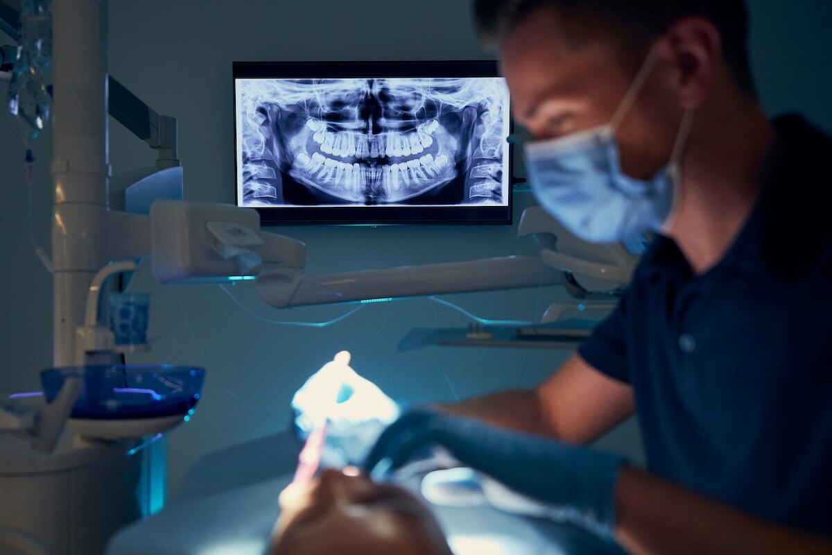 Dentist in face mask treating patient with a large digital panoramic dental X-ray displayed on a wall monitor.