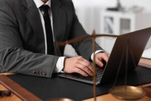 An attorney in a grey suit types on a dark laptop at a wooden desk, representing law firm business operations.