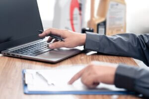 Hands holding pen over laptop screen next to clipboard checklist at desk, reviewing compliance requirements.
