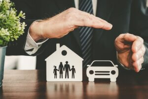 Hands sheltering paper cutouts of a family, house, and car, representing security and insurance protection.