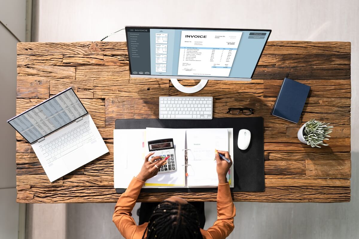 Female Insurance Agent at Desk Top view of person calculating expenses at wooden desk with laptop, monitor displaying invoice, and binder.