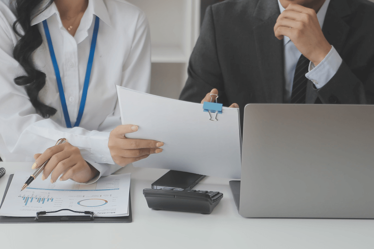 wo professionals seated at a desk reviewing financial reports and paper documents next to a open laptop.